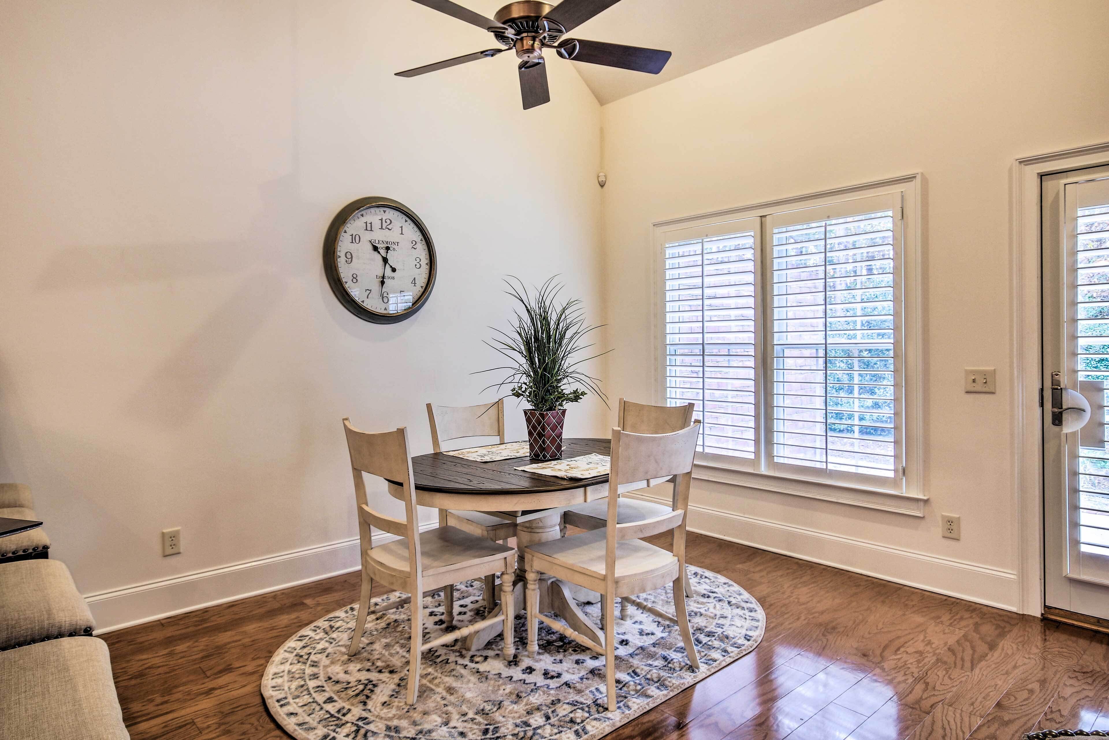 A dining area with a round table, chairs, a clock, a ceiling fan, and a window with blinds.
