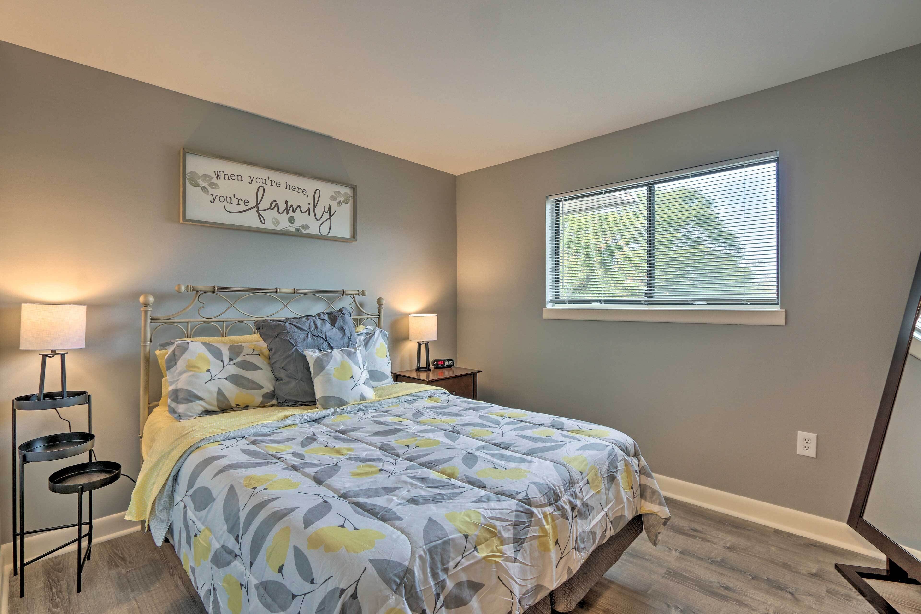 A bedroom with a bed, two bedside tables, a wall clock, a window with blinds, and a motivational quote on the wall.