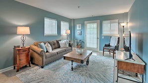 A living room with a sofa, coffee table, side table, and lamp. There are two windows with blinds, a door, and a mirror. - Pools + Sundeck: Beachfront Hilton Head Condo! (Hilton Head Island)