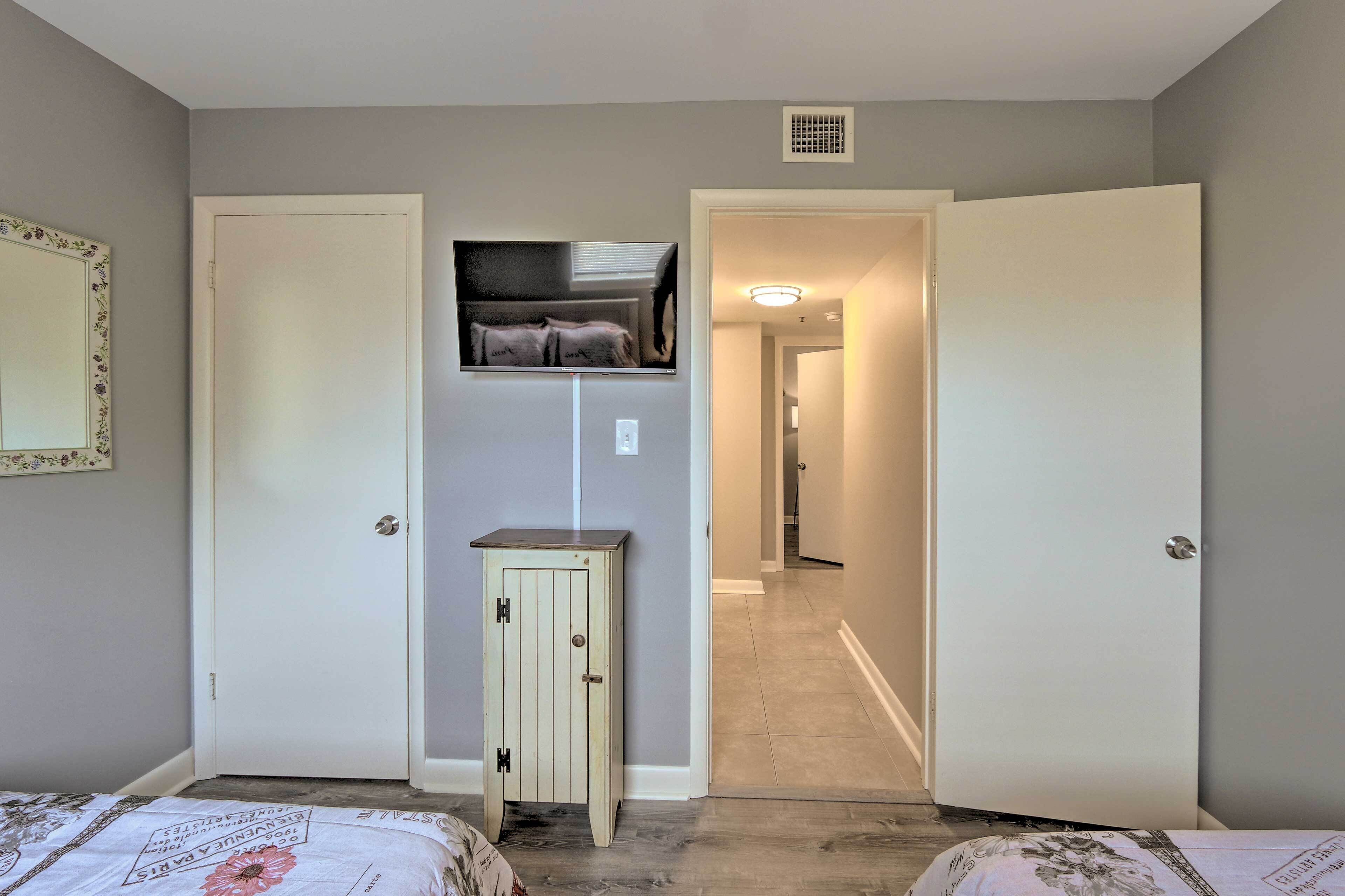 A bedroom with two beds, a small cabinet, a television mounted on the wall, and a hallway leading to another room.