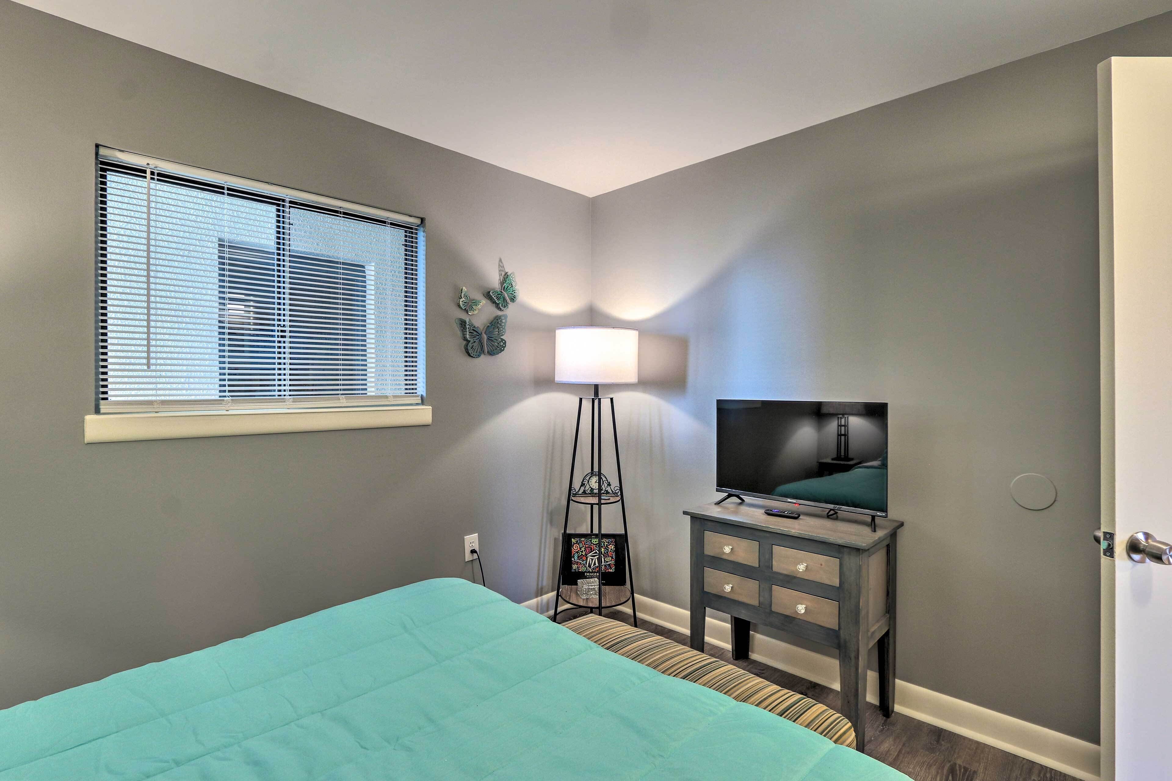 Une chambre à coucher avec un lit, un téléviseur posé sur un meuble, une lampe et une fenêtre avec des stores.