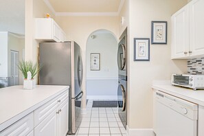 Cocina con armarios blancos, refrigerador de acero inoxidable, microondas y lavavajillas. Hay una planta en maceta sobre la encimera e imágenes enmarcadas en la pared.