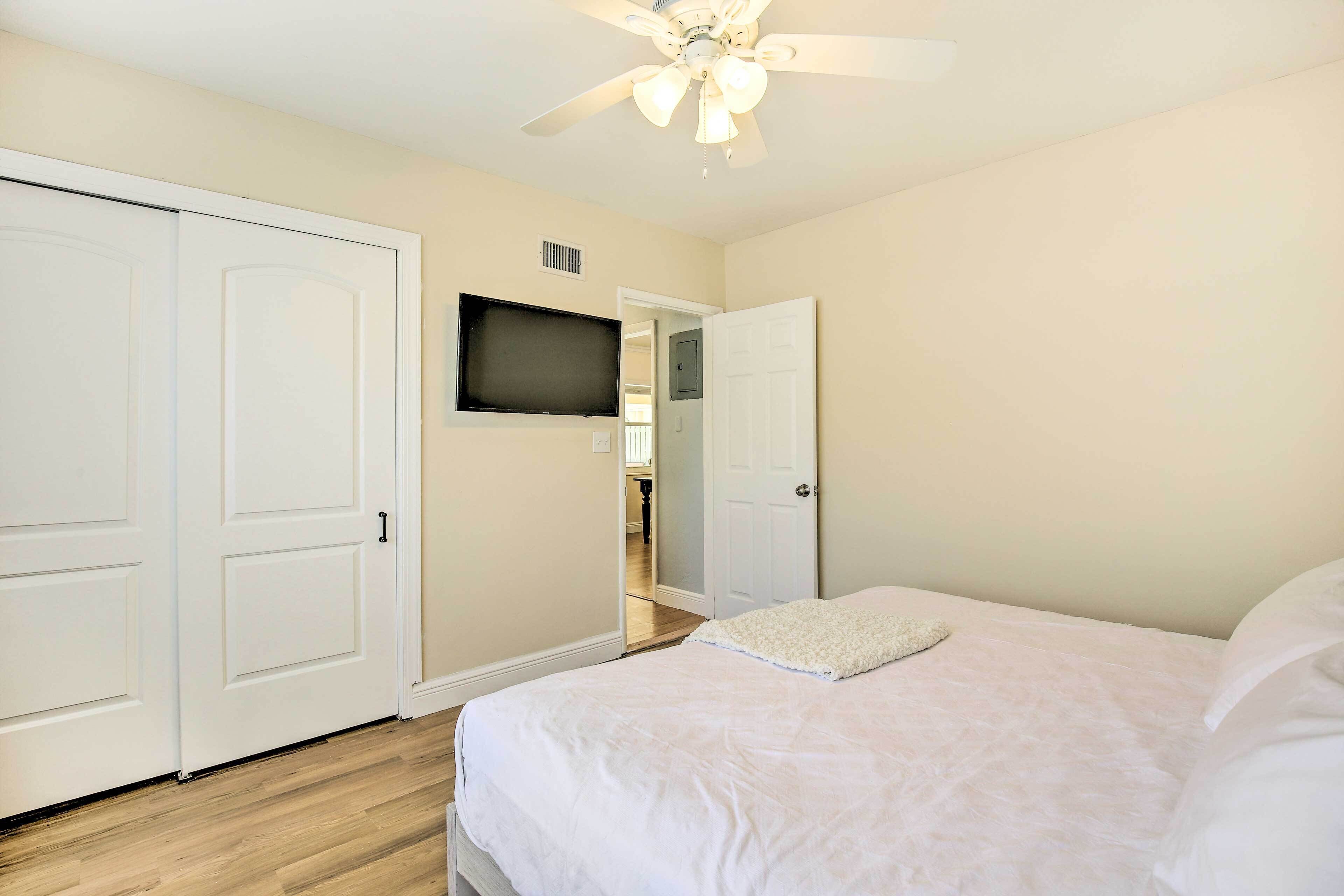 A bedroom with a bed, a ceiling fan, a TV mounted on the wall, and a door leading to another room.