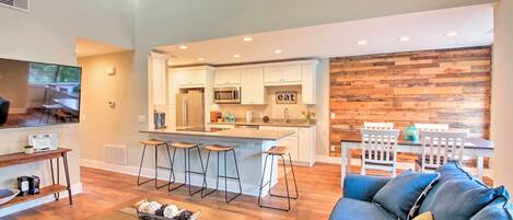 Una sala de estar moderna con una mesa de centro de madera, sofá azul y una cocina con armarios blancos y una pared de acento de madera.