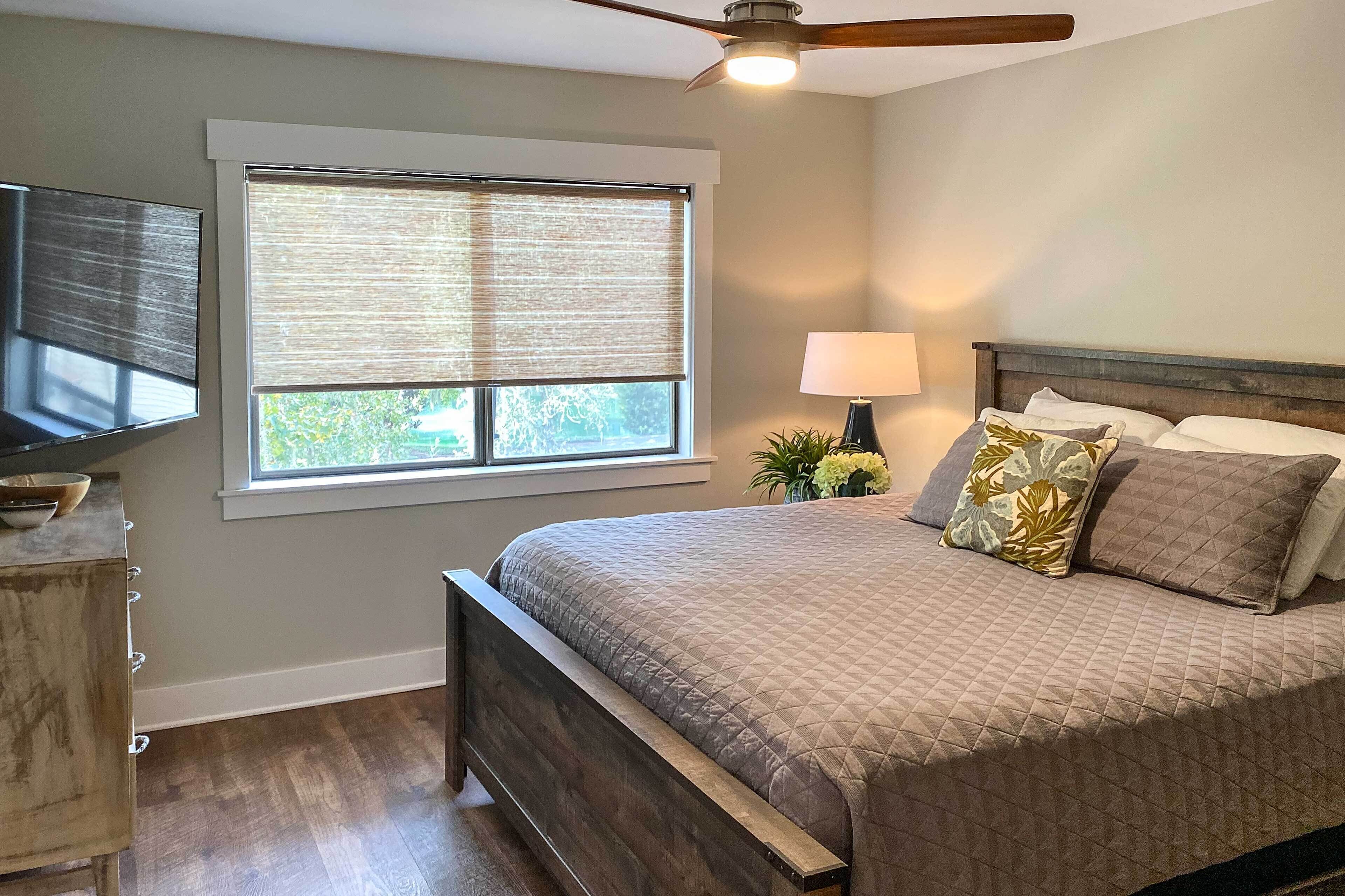 A bedroom with a bed, a ceiling fan, a window with blinds, a bedside table with a lamp, and a TV on a dresser.