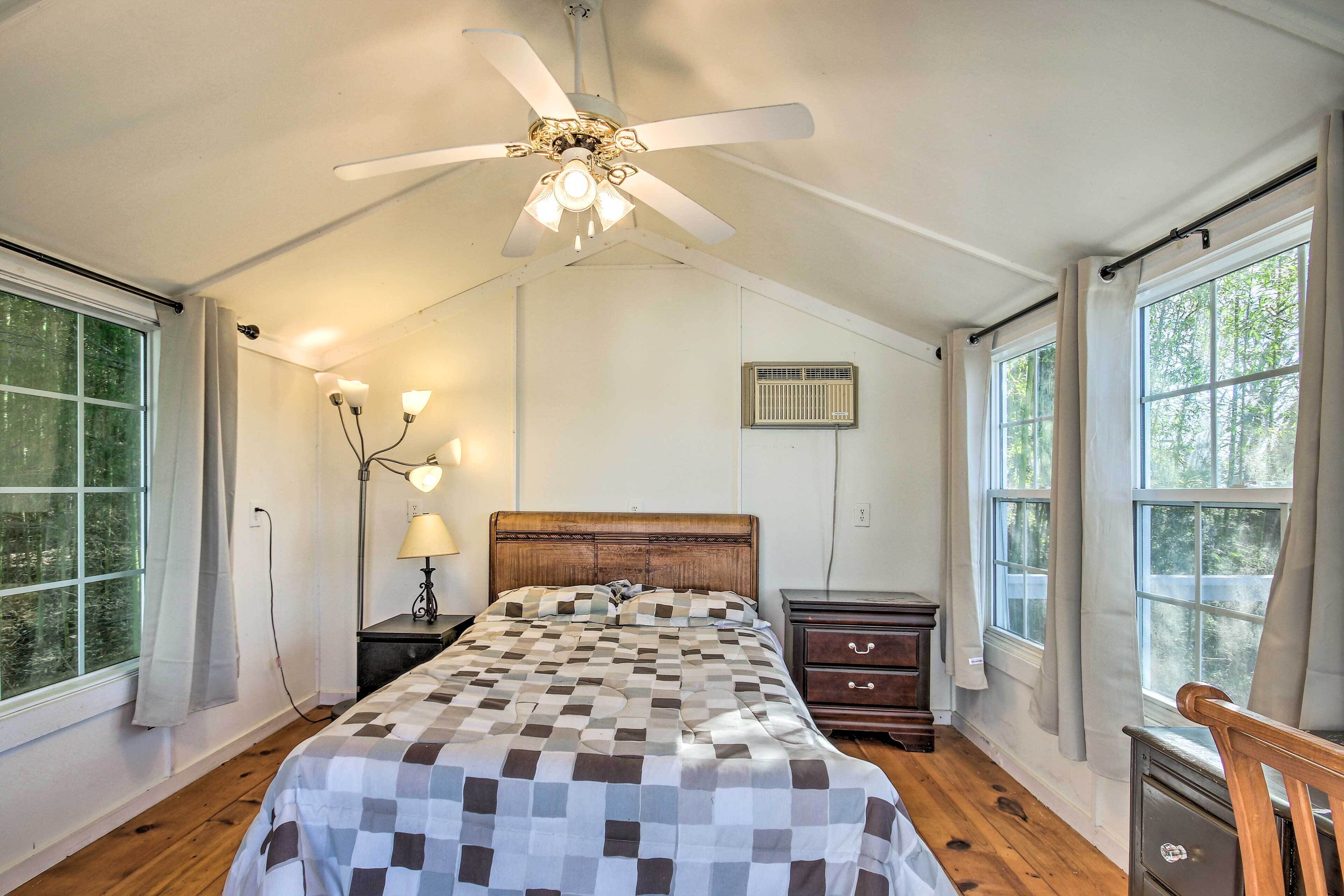 Une chambre à coucher avec un lit en bois, un ventilateur de plafond, une table de chevet, une lampe et une fenêtre avec des rideaux.