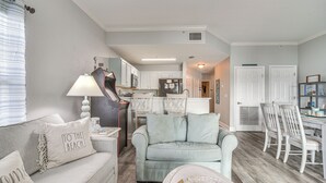 Un salon avec un canapé, une lampe et une petite table. Au fond, on aperçoit une cuisine avec des meubles blancs et un coin repas avec des chaises blanches.