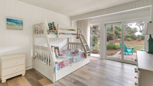 Ein Etagenbett mit farbenfroher Steppdecke, eine weiße Kommode und eine Glastür zum Außenbereich mit Sitzgelegenheiten.
