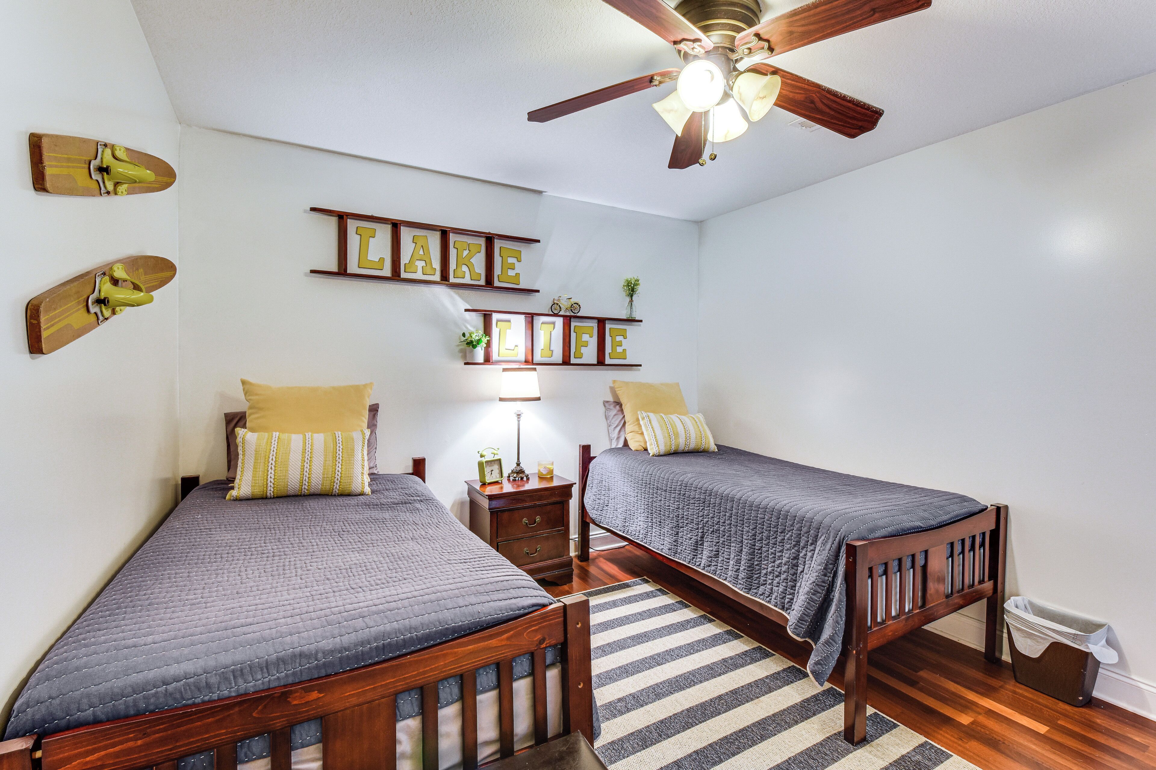 A bedroom with two wooden beds, a ceiling fan, a wall-mounted shelf with framed pictures, and a striped rug.