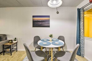 A dining area with a round table, four grey chairs, a small potted plant, and a wall-mounted picture of a sunset over the ocean.