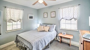 A bedroom with a bed, a nightstand, a desk, a ceiling fan, and two windows with curtains.