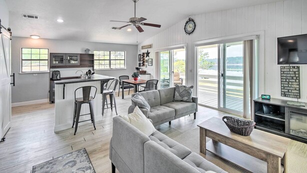 A spacious living area with a grey sofa, a coffee table, and a TV stand. There is a kitchen area with a dining table and chairs, and a large window offering a view of the outdoors.