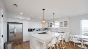 A modern kitchen with white cabinetry, a stainless steel refrigerator, a central island with seating, pendant lighting, and a view through the window.