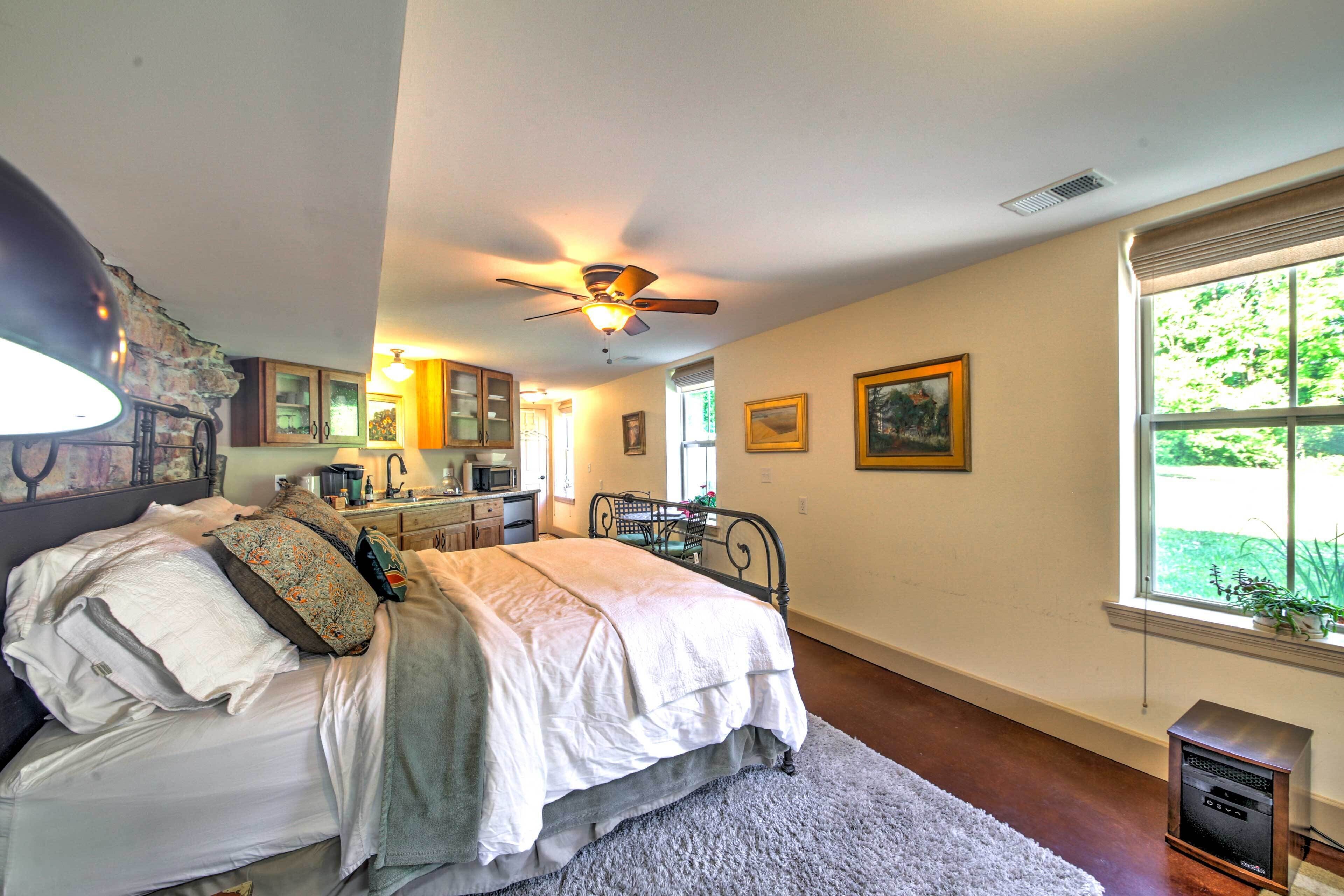 A bedroom with a bed, a ceiling fan, a window with a view of greenery, and a small heater on the floor.