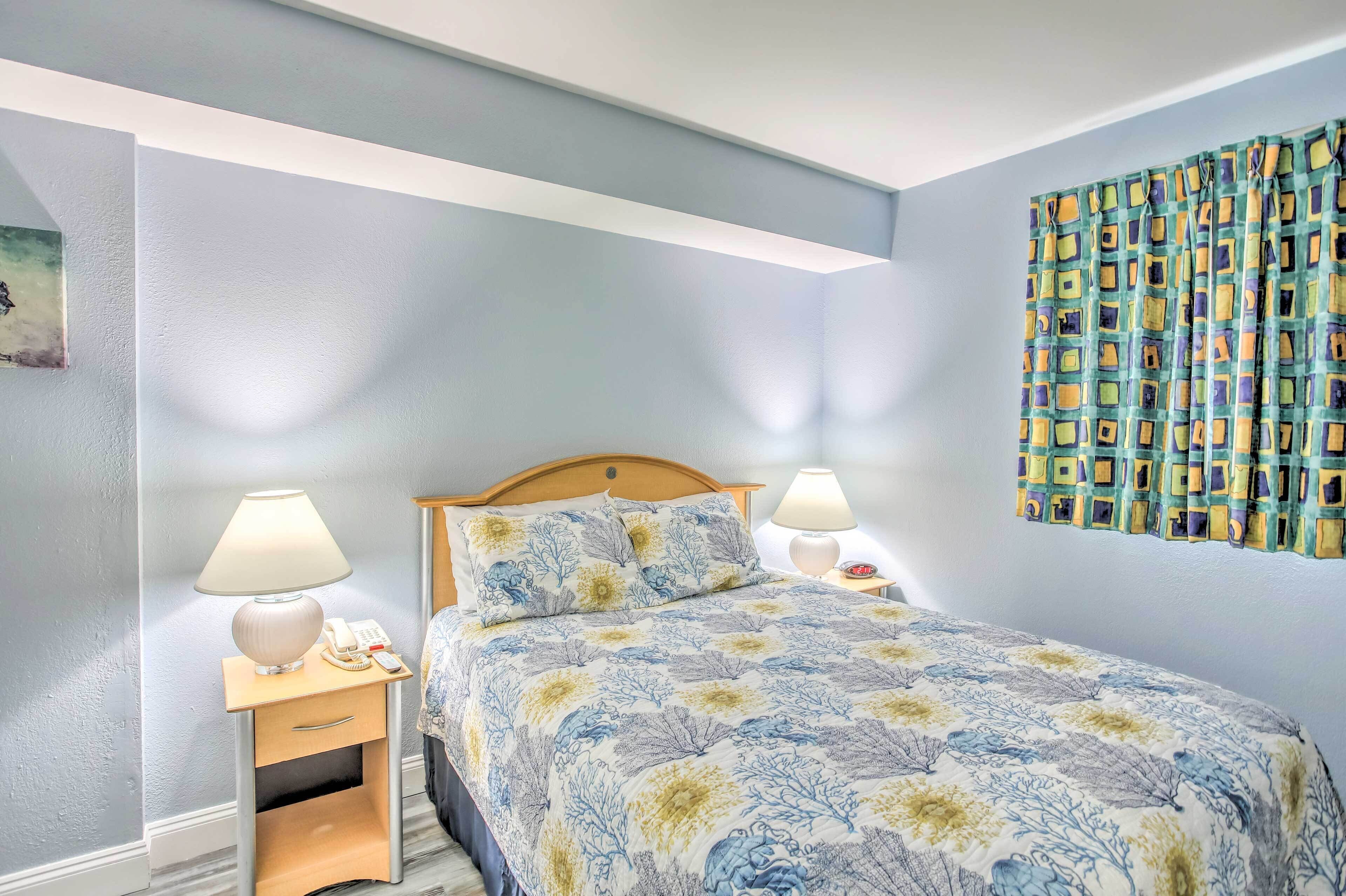 Un dormitorio con una cama, dos mesitas de noche con lámparas, una mesita auxiliar y una cortina estampada.