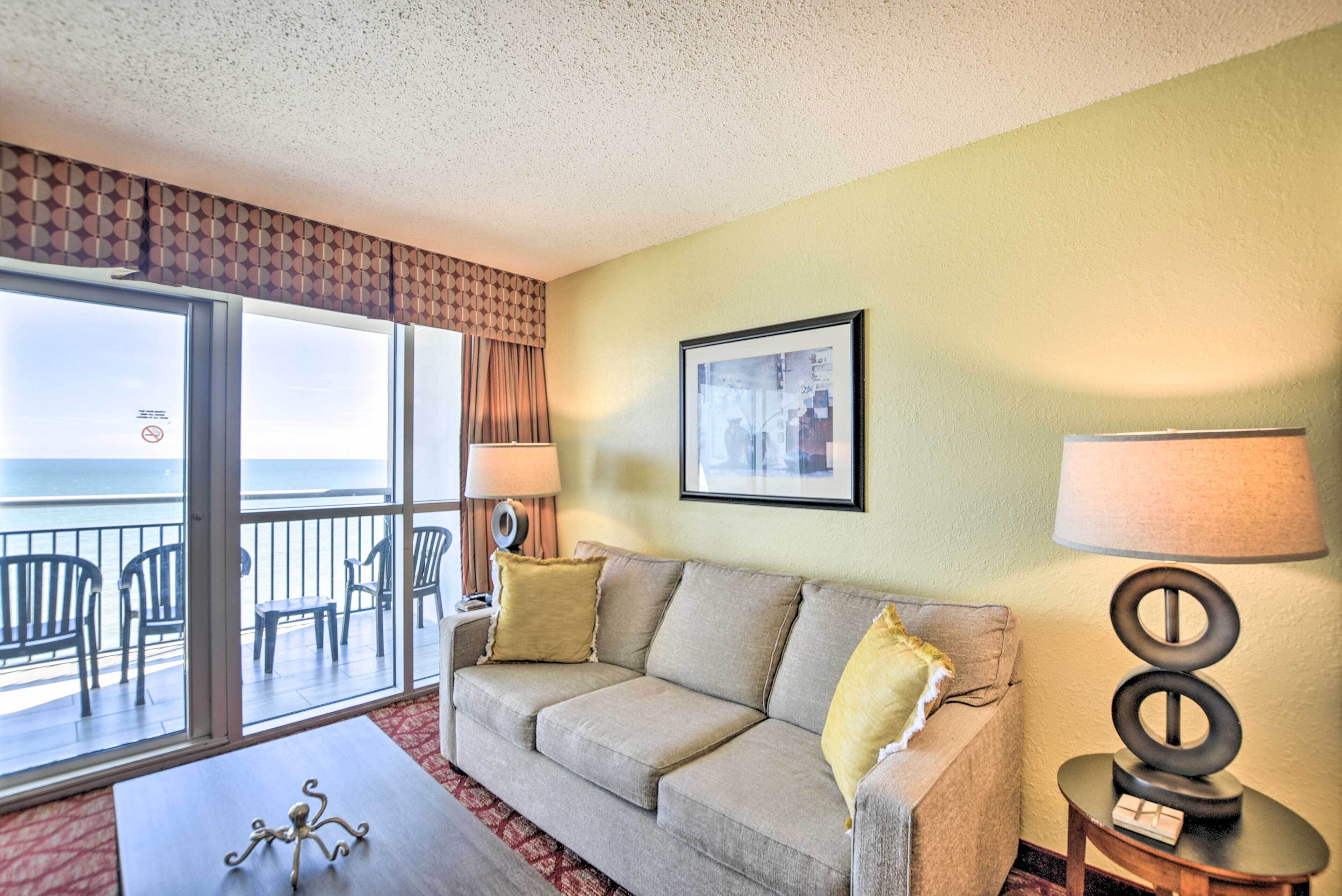 A hotel room with a balcony, a sofa, a coffee table, and a lamp.