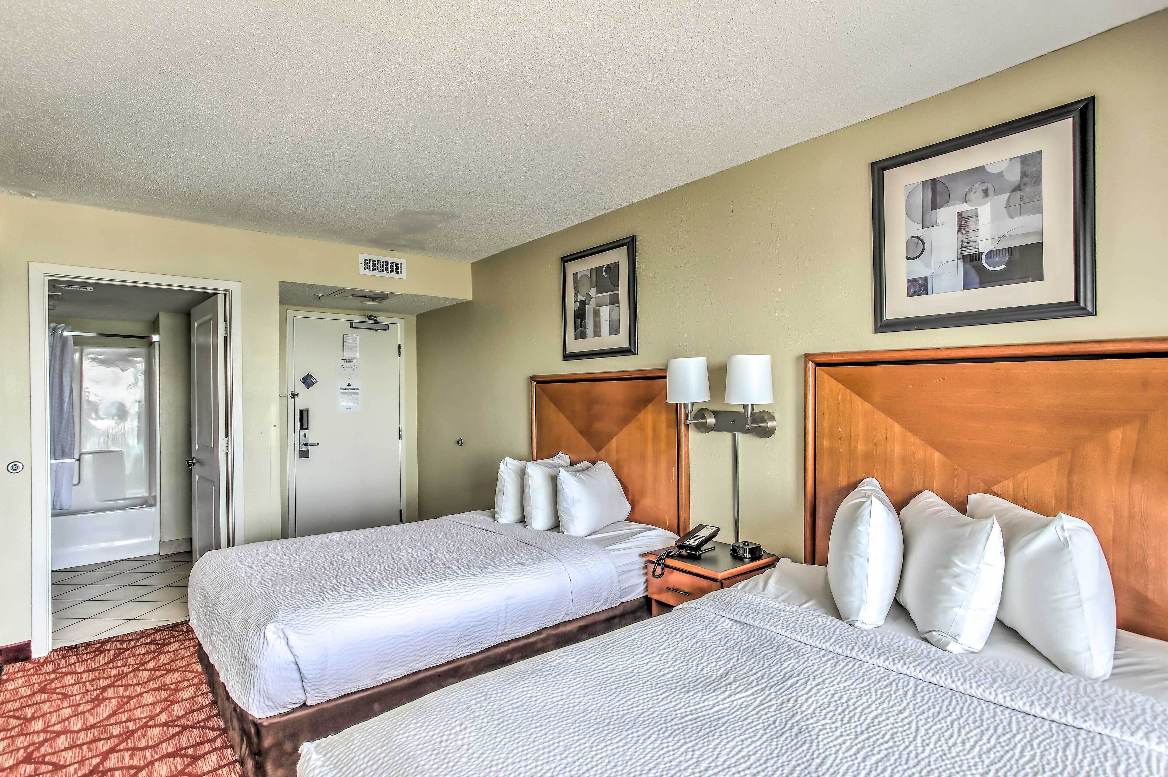 A hotel room with two beds, a wooden headboard, a bedside table with a lamp, and a door leading to a bathroom.