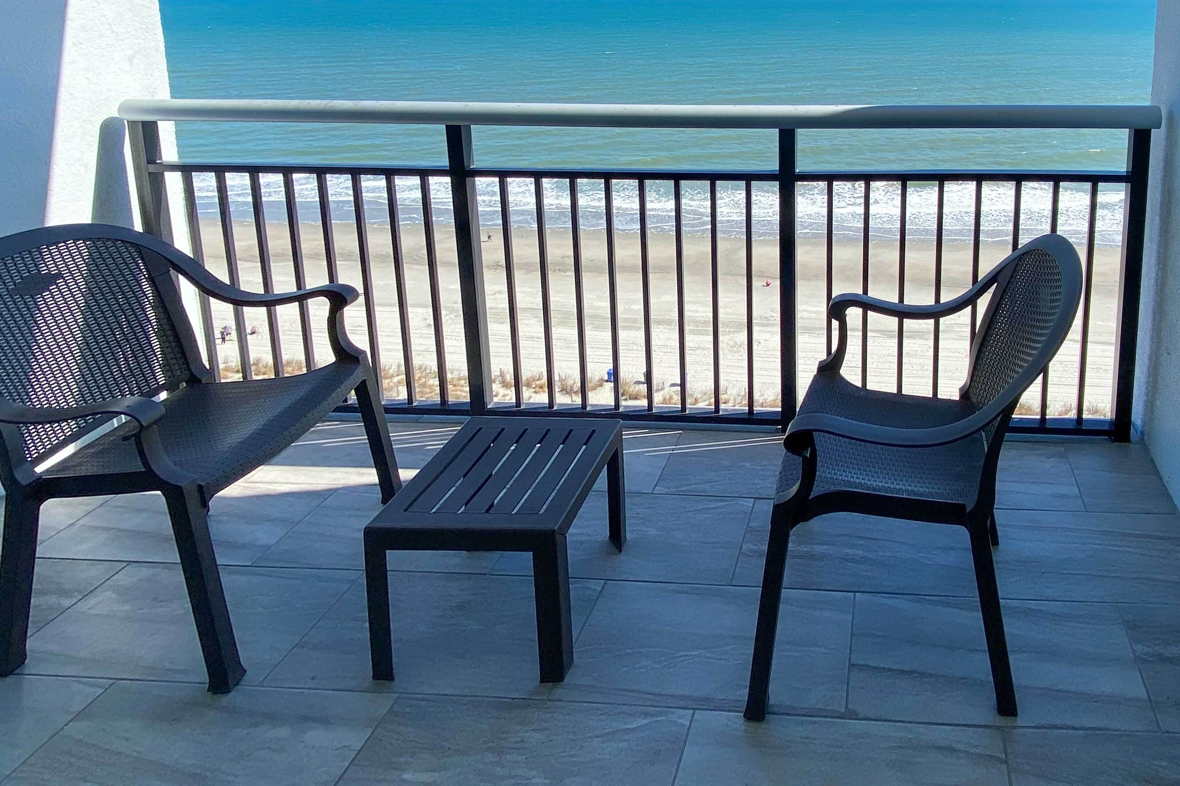 Ein Balkon mit zwei Stühlen und einem kleinen Tisch, der Blick auf einen Strand und einen klaren Ozean bietet.