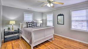 A bedroom with a white bed, wooden flooring, a ceiling fan, and two windows with blinds.