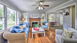 A spacious living room with a fireplace, large windows, and a ceiling fan.