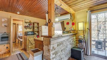 A rustic cabin interior with a stone bar, wooden beams, a fireplace, and a large window.