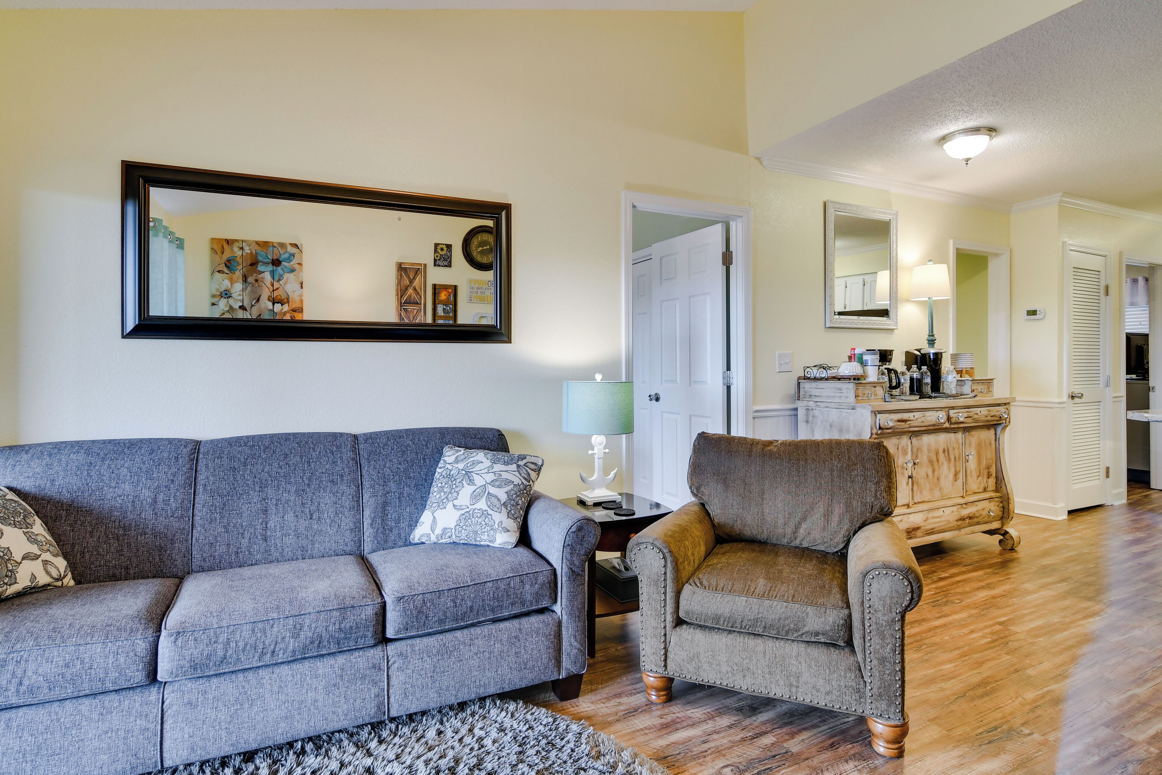 A living room with a gray sofa, a brown armchair, and a coffee table. There is a painting on the wall and a mirror above the sofa.