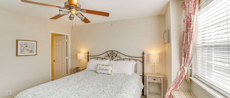 A bedroom with a bed, a ceiling fan, bedside lamps, a window with blinds, and a door.