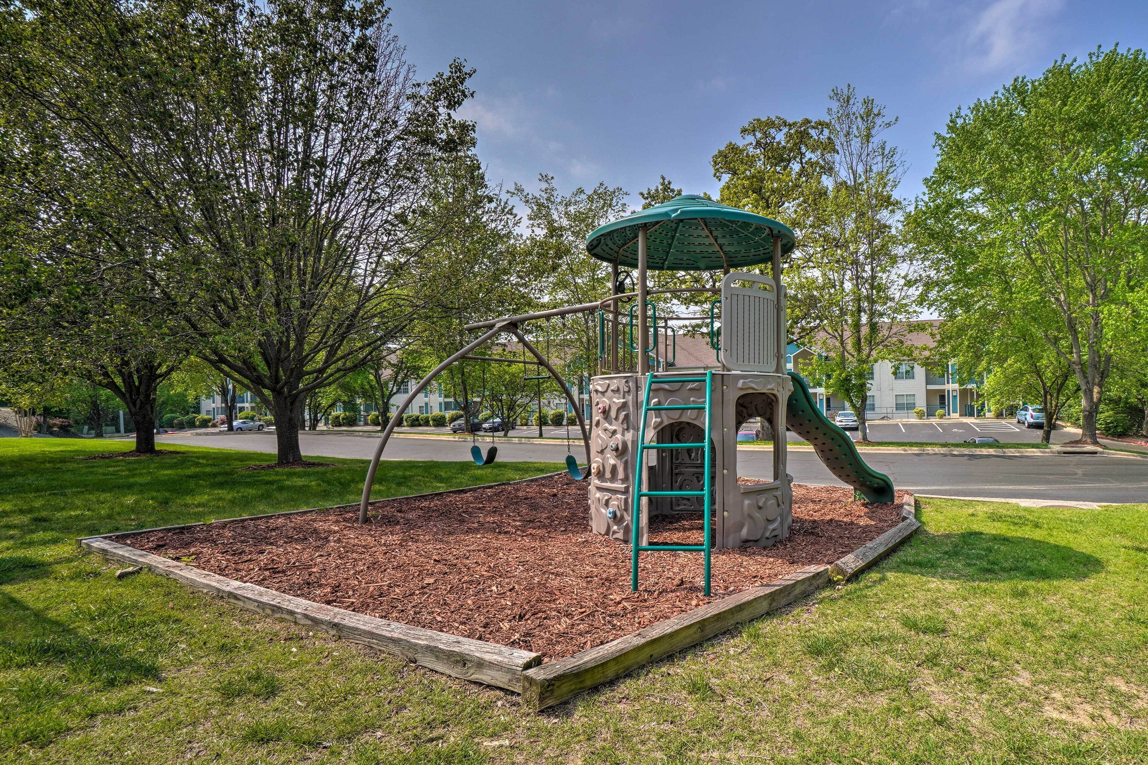 A playground with a slide and swings, surrounded by grass and trees.