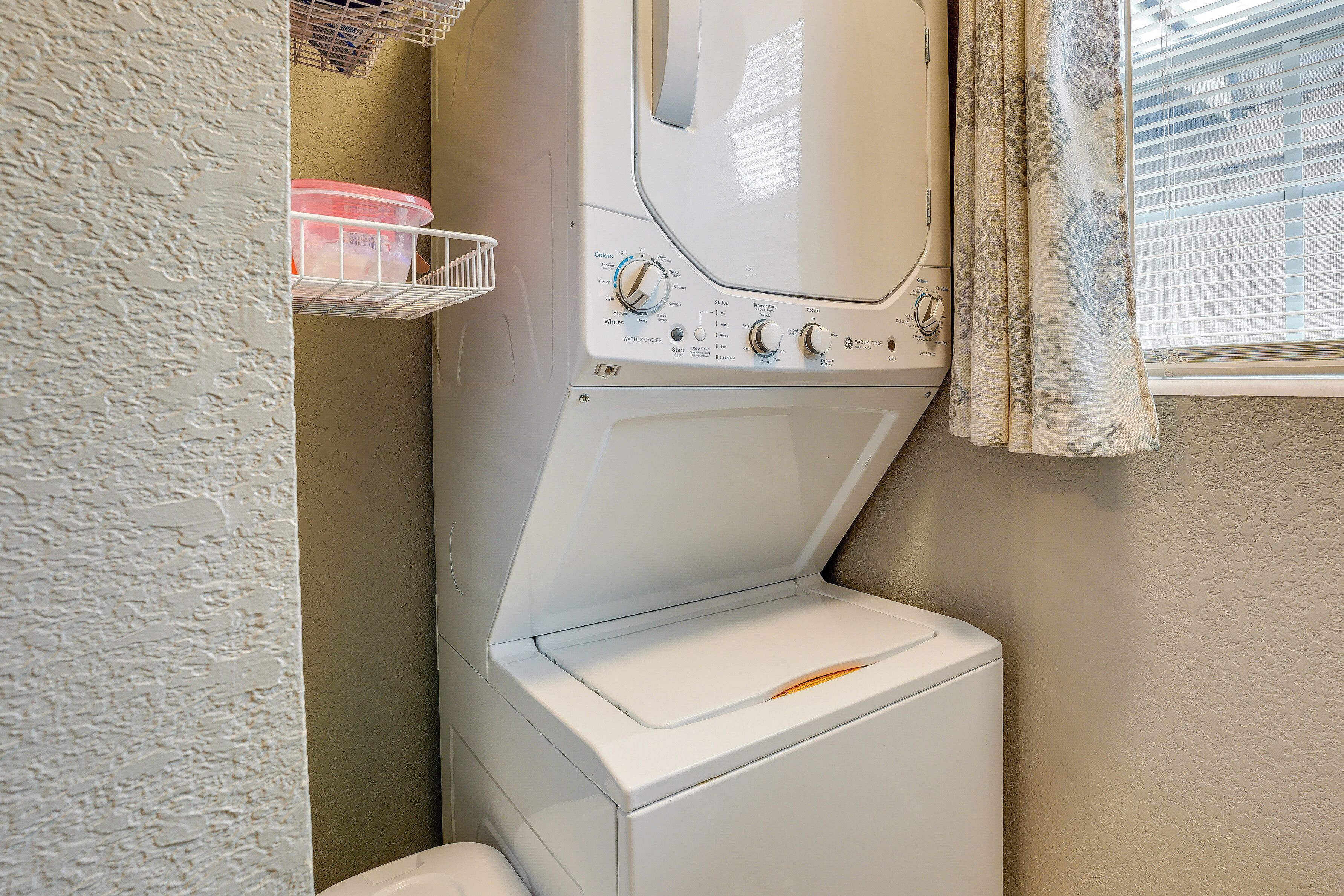 A front-loading washing machine with a control panel, a white wall, and a window with curtains.