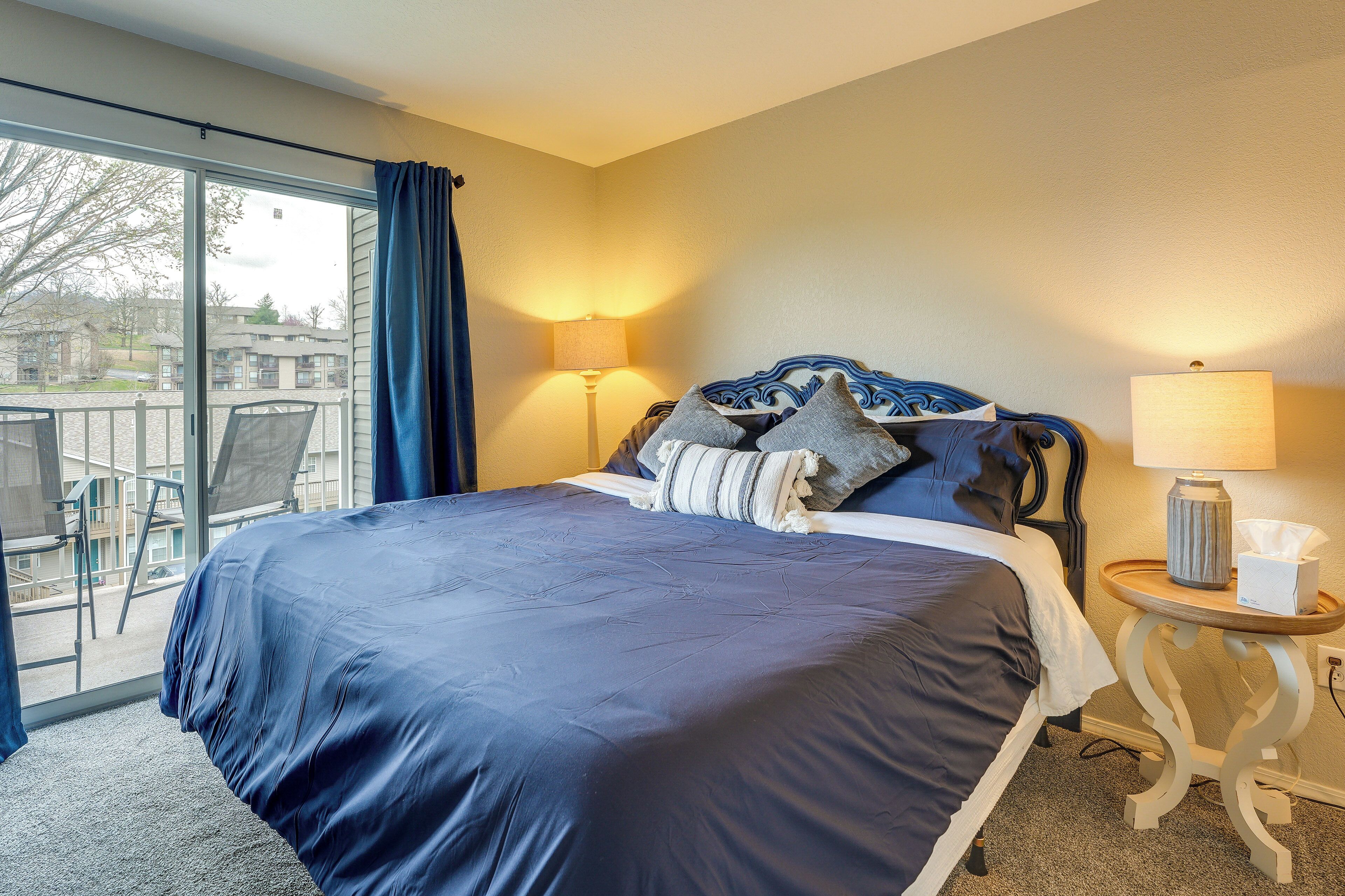 A hotel room with a large bed, two bedside lamps, a small table with a tissue box, and a view of the outside balcony.