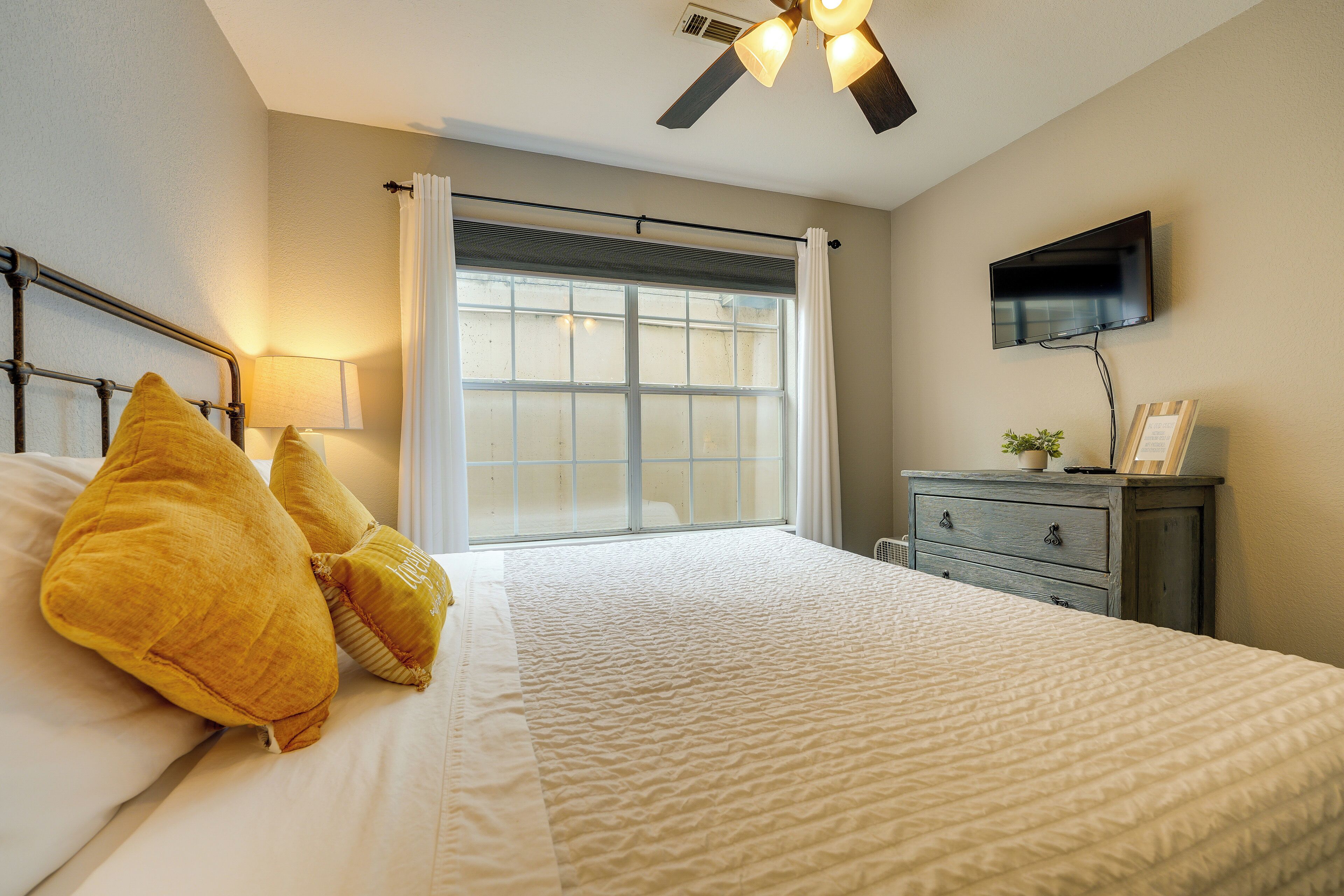 Un dormitorio con cama, mesita de noche, cómoda, ventilador de techo, ventana con cortinas y televisor de pantalla plana montado en la pared.