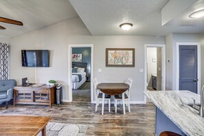 A living room with a wooden coffee table, a gray sofa, and a TV mounted on the wall. There is a dining area with a wooden table and chairs, and a kitchen area with a marble countertop and sink.