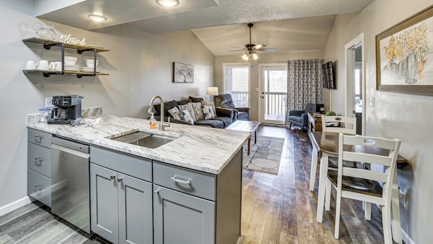 A modern kitchen with a marble countertop, grey cabinets, and a built-in dishwasher. There is a dining area with a table and chairs, and a living room with a sofa and a ceiling fan. - Family-friendly Condo: 1 Mi to Silver Dollar City! (Branson West)