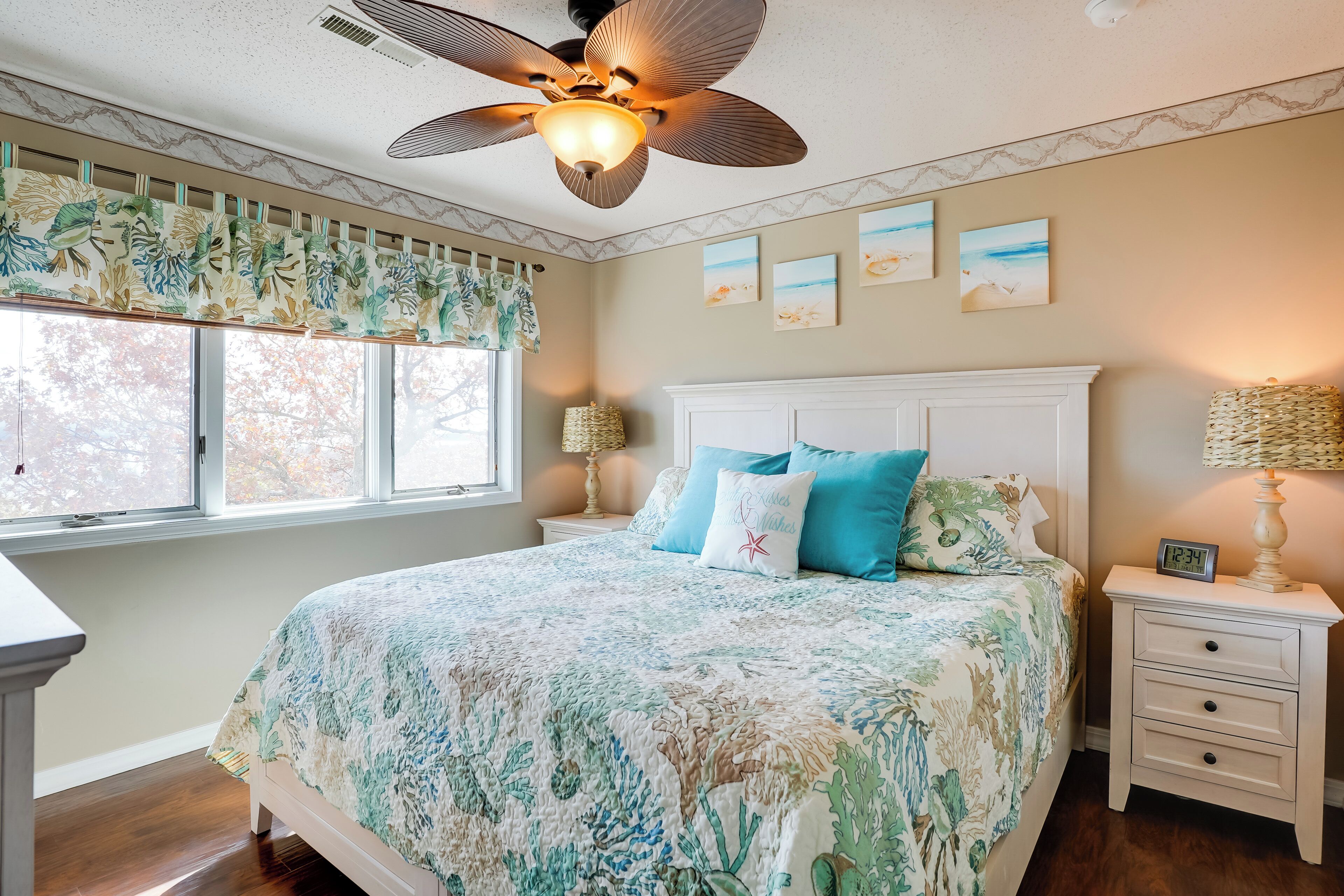 A bedroom with a bed, a nightstand, a ceiling fan, a window with curtains, and wall art.