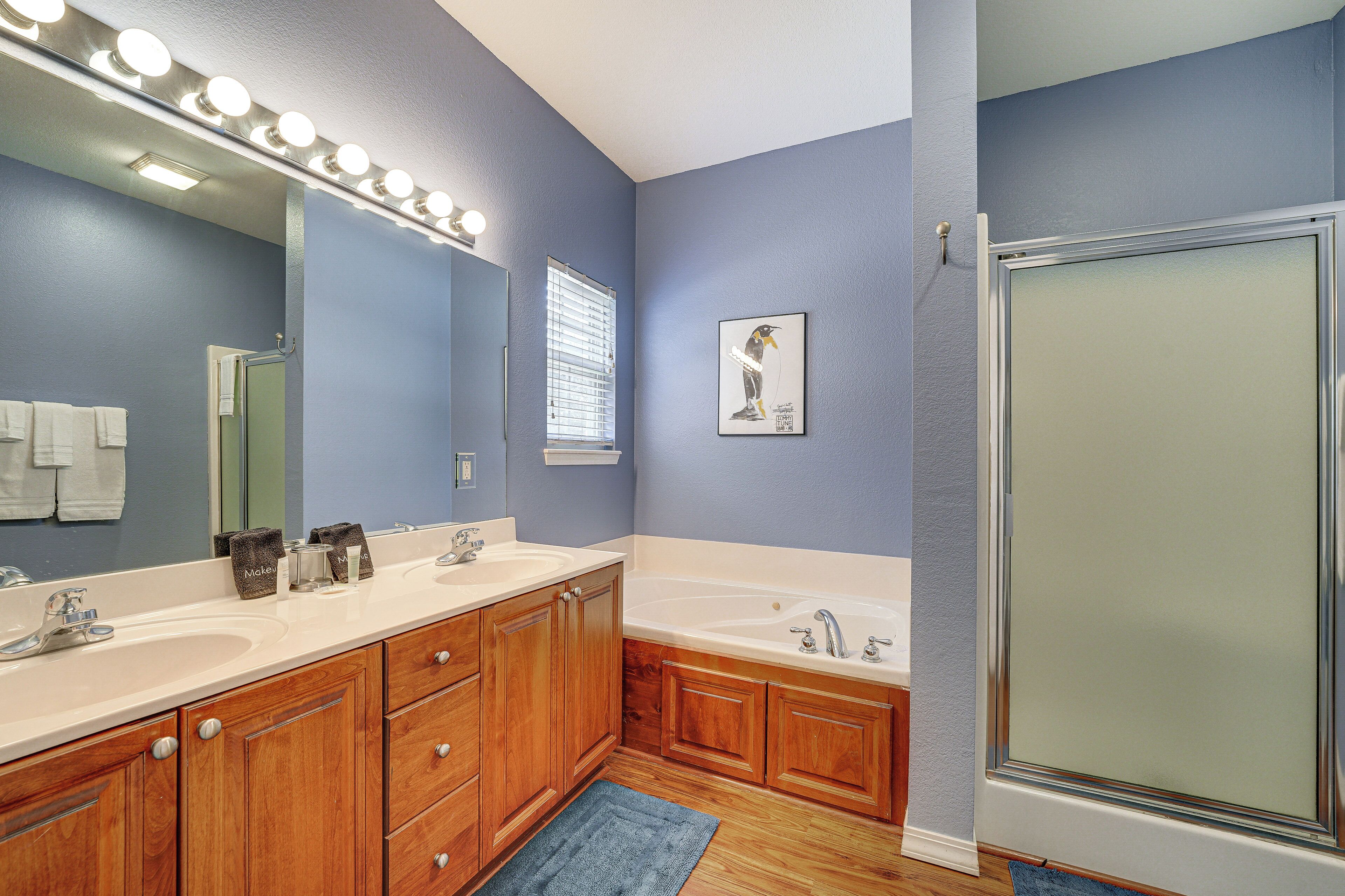 A bathroom with two sinks, wooden cabinets, a large mirror with lights, a shower, and a framed picture on the wall.