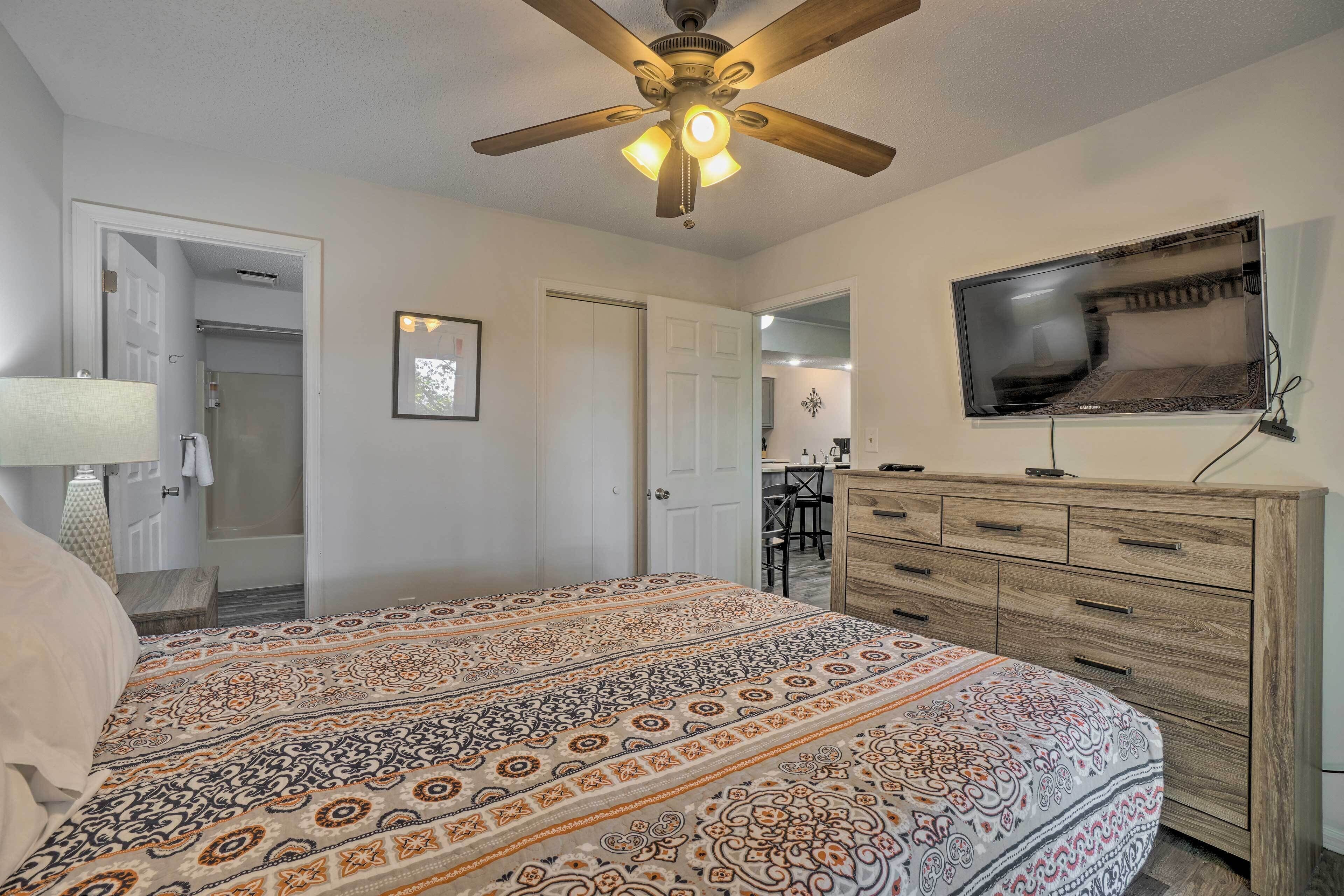 A bedroom with a patterned bedspread, a ceiling fan, a TV mounted on the wall, and a wooden dresser.