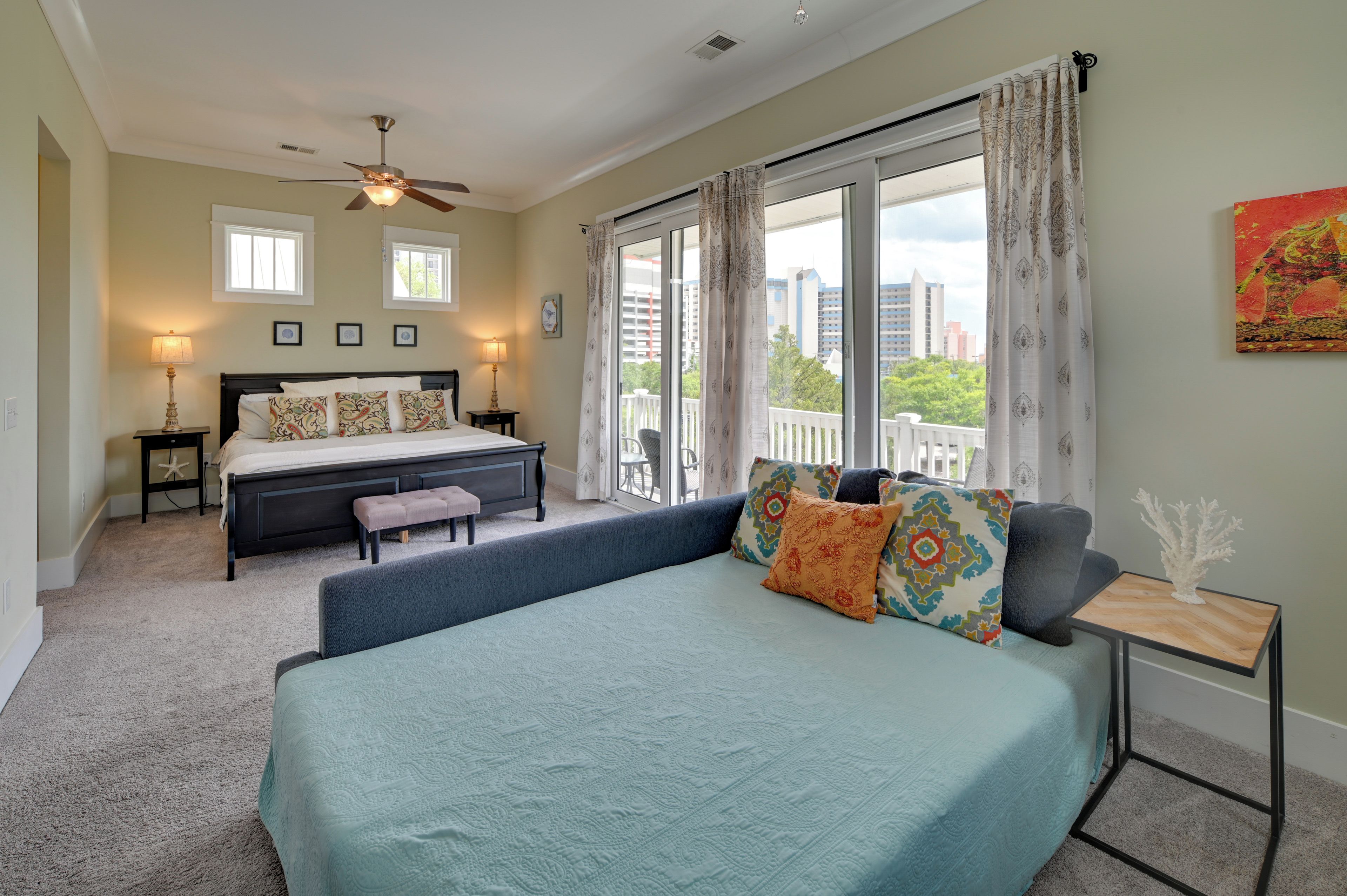 A bedroom with a large bed, a bench, a bedside table, a ceiling fan, and a view of the city through large windows.