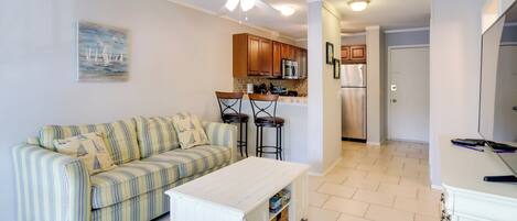 A living room with a striped sofa, a white coffee table, and a green armchair. There is a kitchen area with wooden cabinets and a refrigerator.