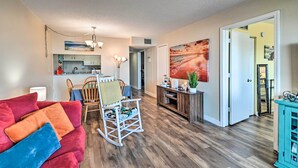A living room with a red sofa, wooden flooring, a rocking chair, a dining area with a table and chairs, and a kitchen in the background. - Bright Myrtle Beach Condo w/ Private Balcony! (Myrtle Beach)
