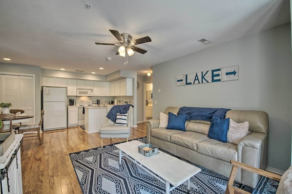 A modern living room with a beige sofa, a coffee table, and a patterned rug. There is a kitchen area with white cabinets and a microwave. A ceiling fan is visible, and a sign on the wall reads "LAKE".