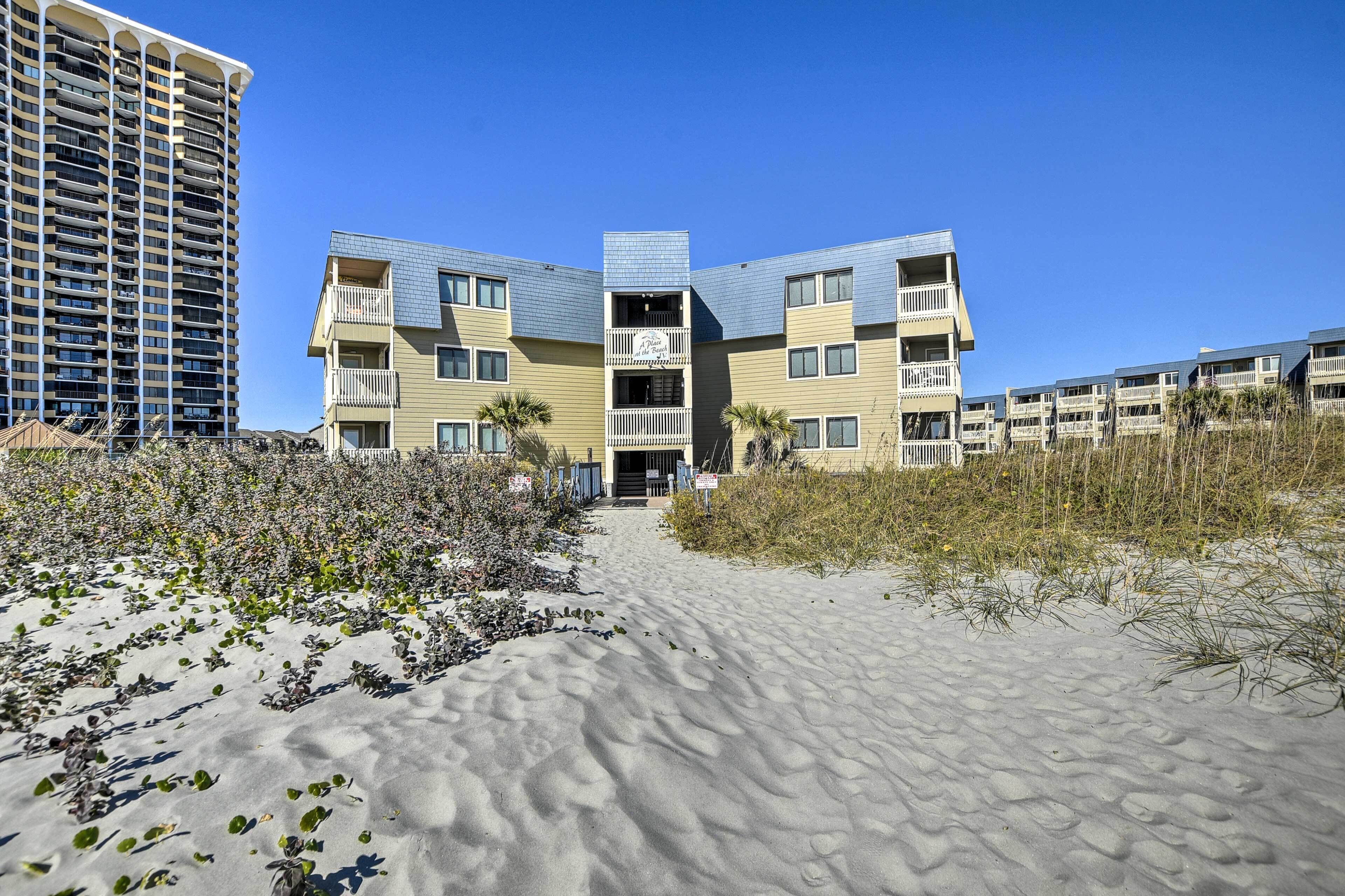 Ein Strandhotel mit einem sandigen Weg zum Eingang, mehreren Balkonen und einem hohen Gebäude im Hintergrund.