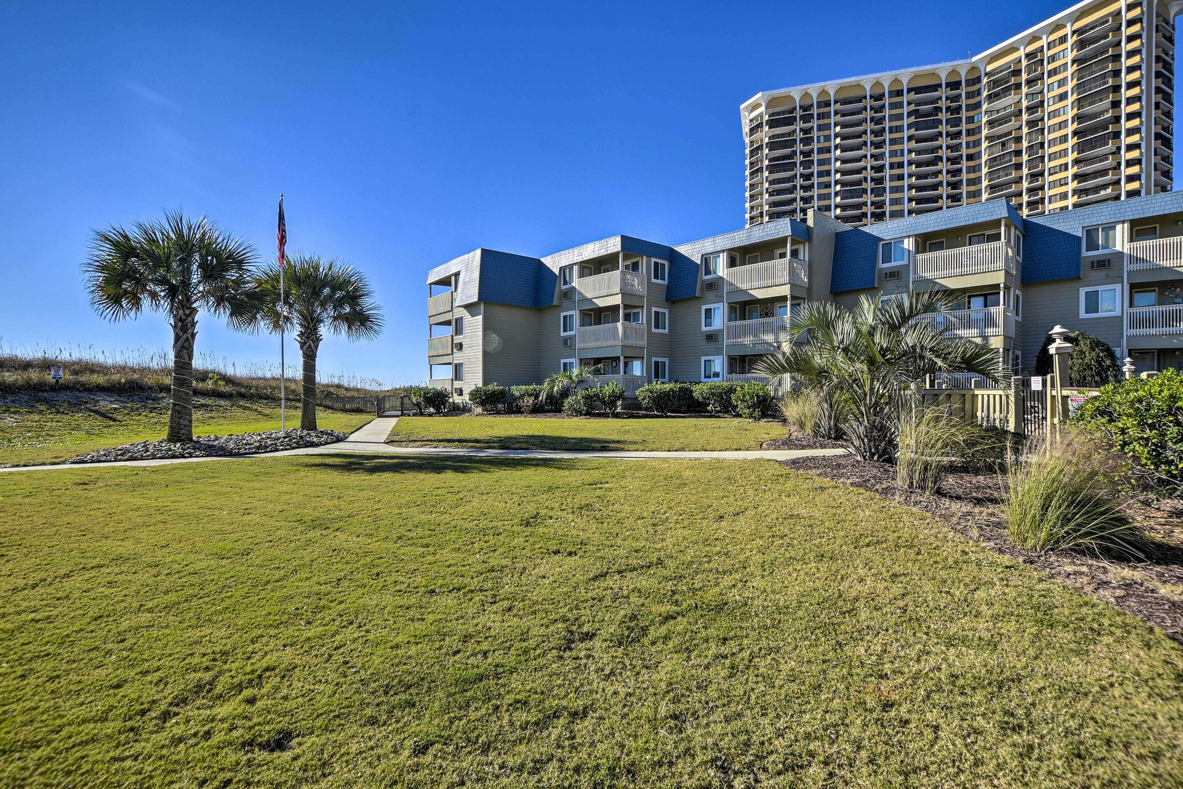 A multi-story hotel with balconies, a well-maintained lawn, and palm trees.