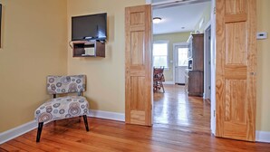 A room with a wooden floor, a patterned chair, a television mounted on the wall, and a kitchen area visible through an open door. - Charming Berger Apt on 42-acre Farm w/ Pool Access (Berger)
