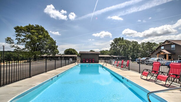 A rectangular swimming pool with a clear blue water surface, surrounded by a black metal fence. There are red lounge chairs along the poolside, and a building with a dark roof and windows is visible in the background. - Cozy Resort Cabin: Walk to Lake + Pool! (Lampe)