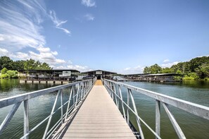 A wooden pier leading to a covered boat launch area with a metal railing, surrounded by water and greenery. - Cozy Resort Cabin: Walk to Lake + Pool! (Lampe)