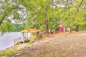 A lakeside cabin with a dock, a covered seating area, and a small building with a pink exterior. - Cheery Swansea Home on Calmont Pond w/ Dock! (Swansea)