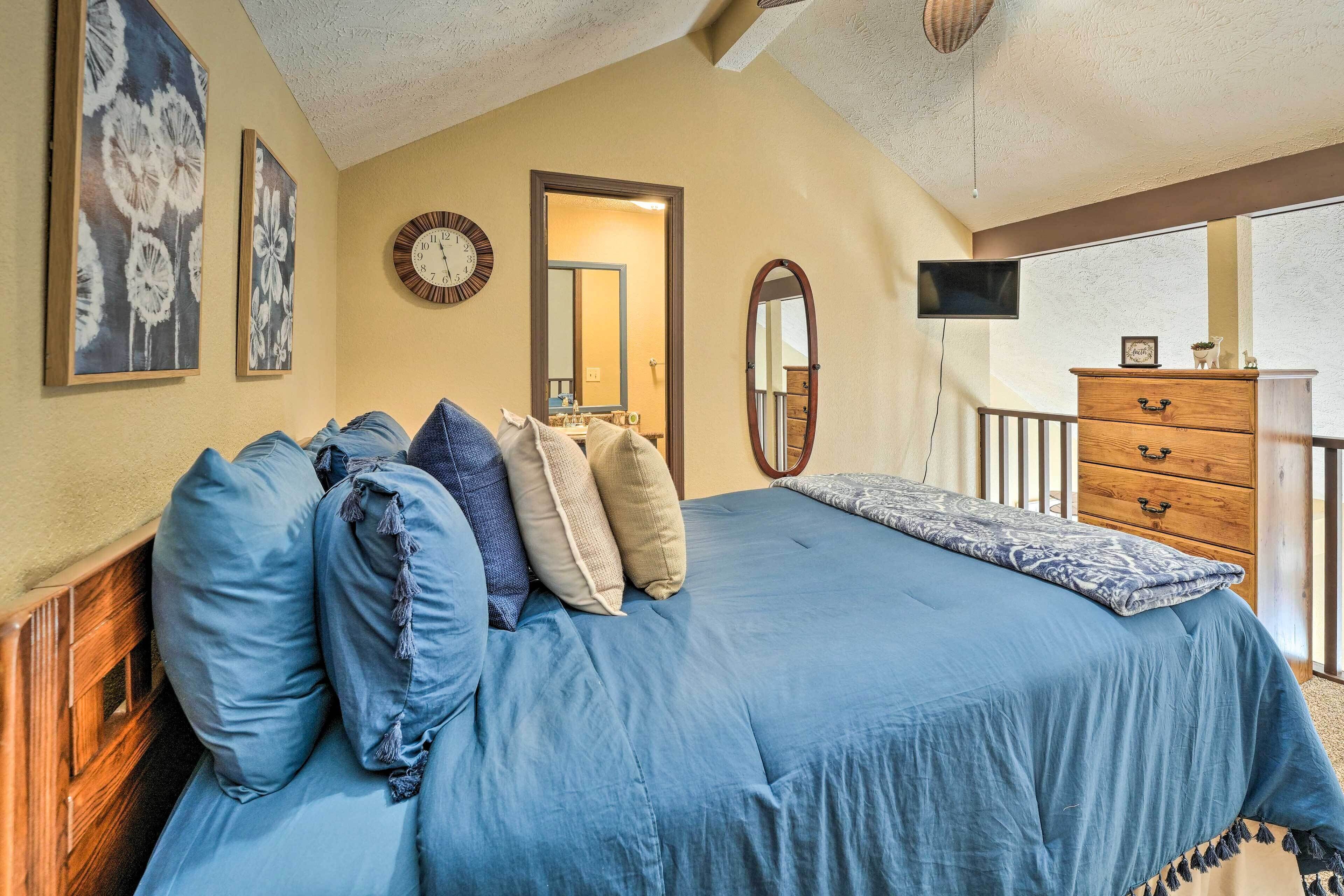 A bedroom with a large bed, a wooden dresser, a mirror, and a TV on the wall.