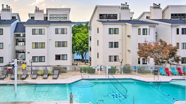 A swimming pool surrounded by white apartment buildings with balconies, a fenced deck area with lounge chairs, and a clear sky.