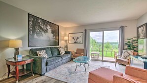 A living room with a sofa, a glass coffee table, a red patterned ottoman, a wooden side table, and a lamp. There is a large painting on the wall, a framed picture of a bull, and a view of greenery through the glass door. - Cheerful Condo: Patio, 2 Mi to Branson Strip! (Branson)