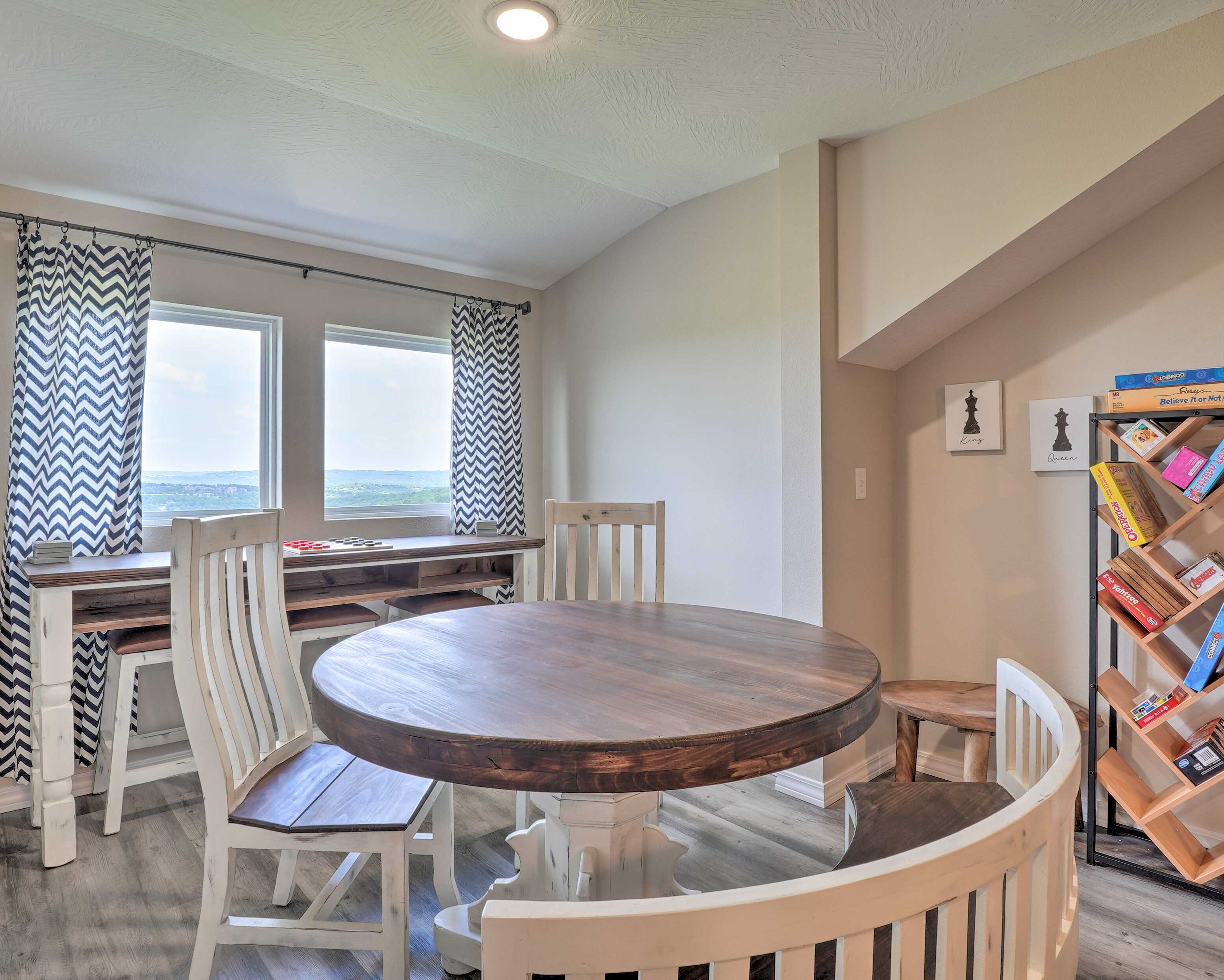 A dining area with a round wooden table, white chairs, a window with a view, and a bookshelf with books.